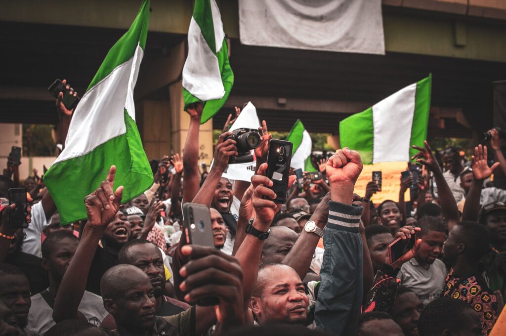 A dense crowd beneath a flyover raises Nigerian flags, fists and phone cameras.