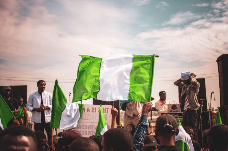 A large Nigerian flag is waved above a crowd facing a stage, with a placard reading This is a new Nigeria, #EndSARS #EndSWAT, visible below.