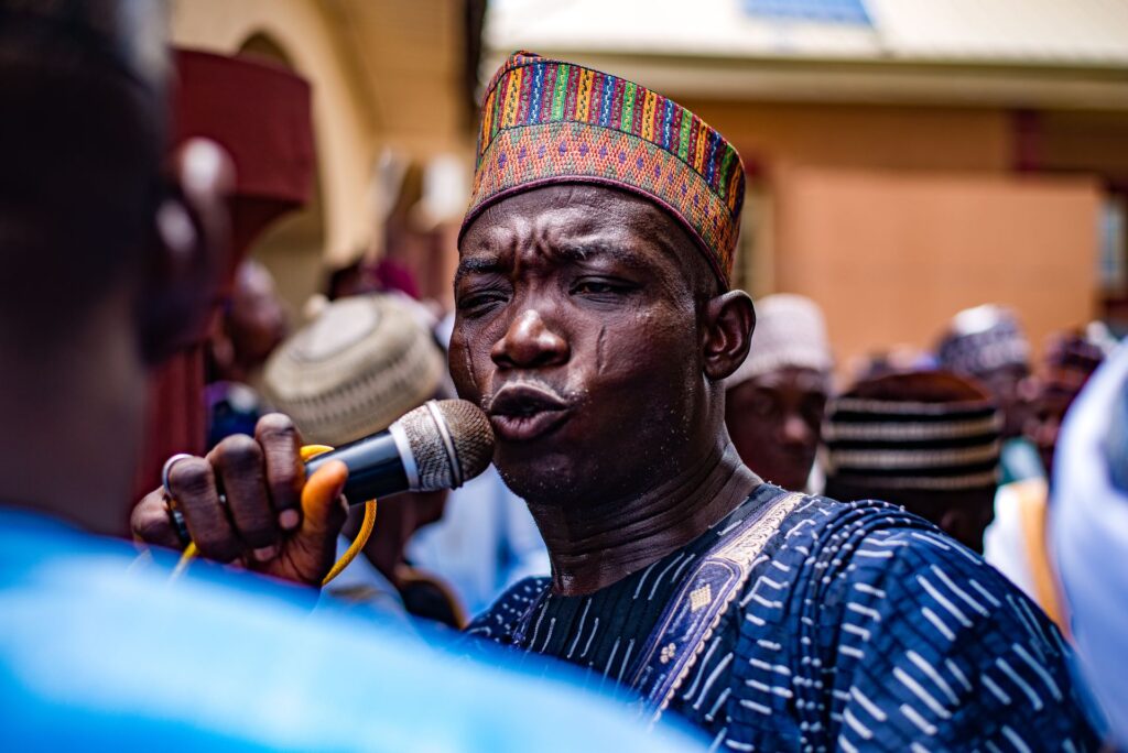 A man in a brightly patterned cap speaks into a microphone, with rows of men in embroidered caps behind him.