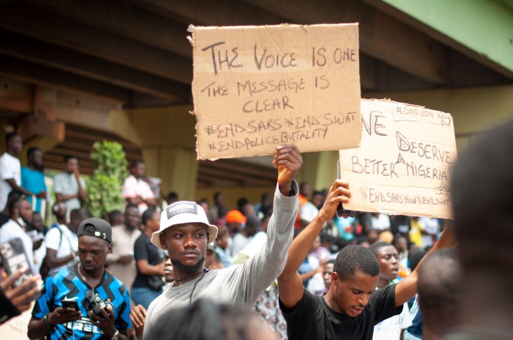 A young man in a white bucket hat holds a cardboard sign reading The voice is one, the message is clear, #EndSARS #EndSWAT #EndPoliceBrutality; a second sign beside it reads We deserve a better Nigeria.