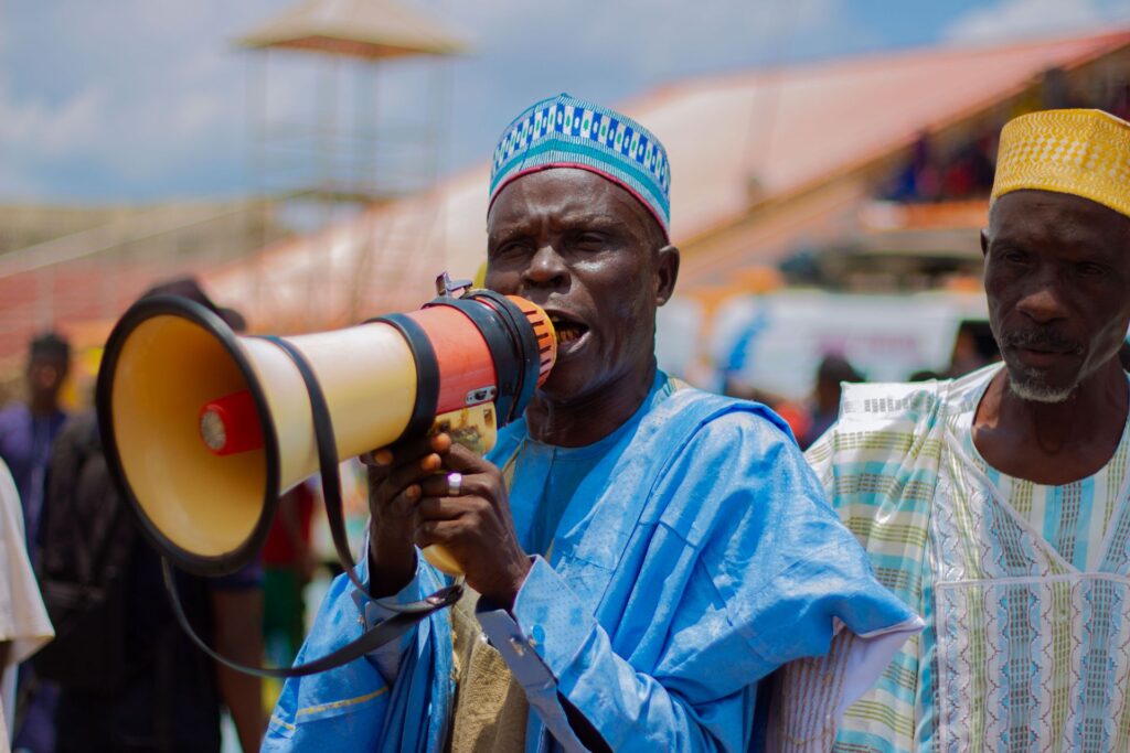 An older man in a light blue kaftan and embroidered cap speaks through a large megaphone at an outdoor gathering, another elder standing beside him.