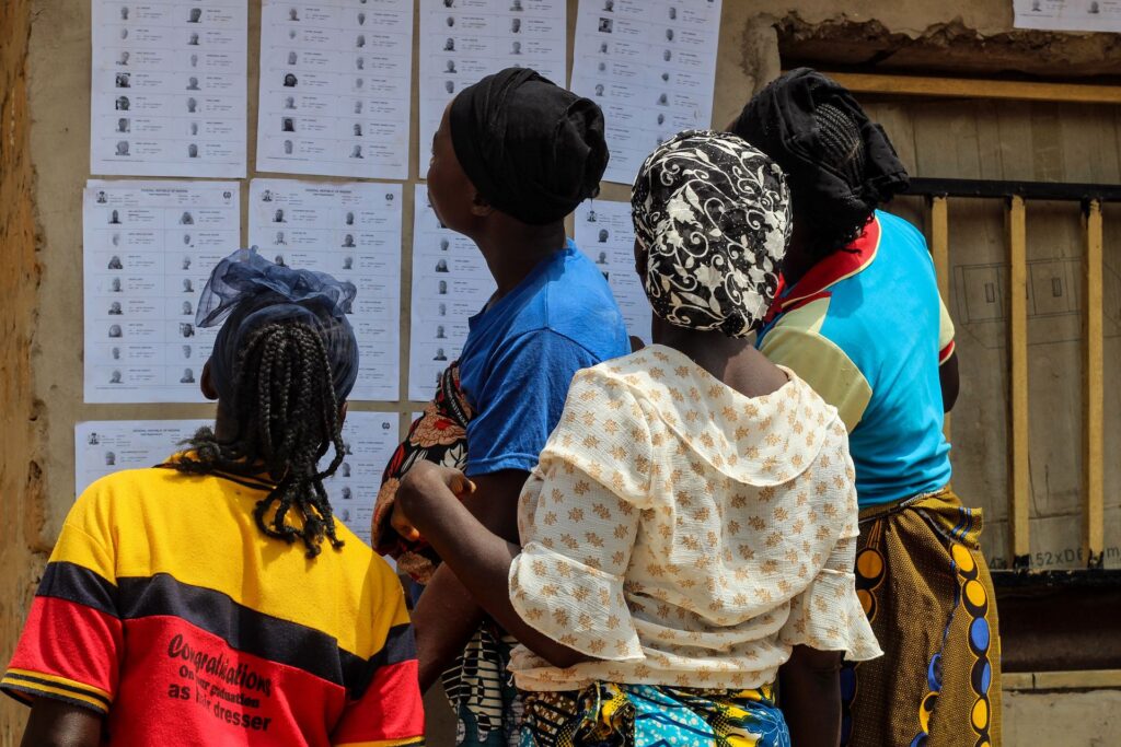 Four women stand with their backs to the camera reading voter registration lists posted on a wall outside a polling unit.