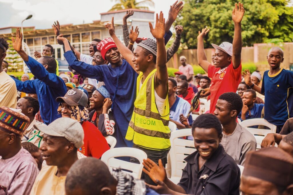 A seated outdoor crowd raises their hands and cheers, with a man in a high-visibility vest marked SIECOM standing among them.