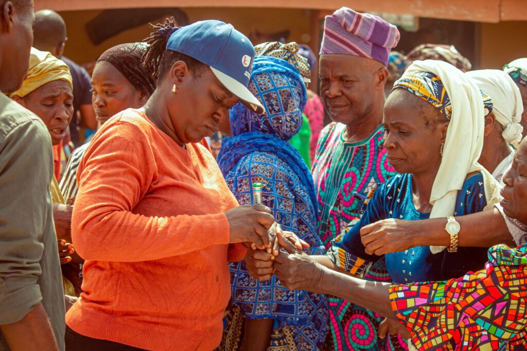 An official in an orange sweater marks a voter's finger with ink at a polling unit, surrounded by men and women waiting their turn.