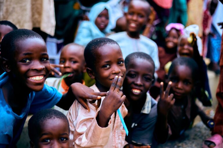 Smiling children lean towards the camera outdoors, several making peace signs with their hands.