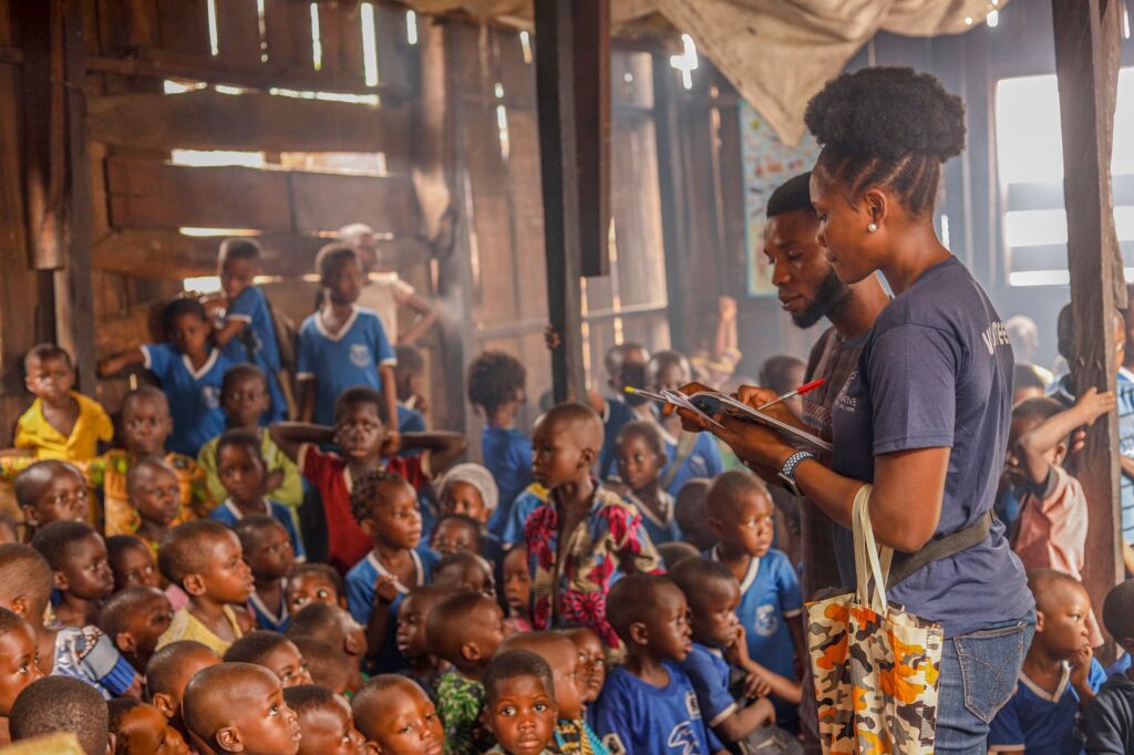 Two A-Goal Initiative volunteers make notes on a clipboard while addressing a packed classroom of primary school pupils.