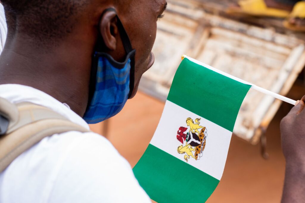 A man in a face mask, seen over his shoulder, holds a small Nigerian flag bearing the national coat of arms.