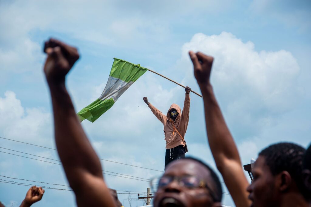 A masked protester stands above the crowd with arms outstretched, waving a worn Nigerian flag on a stick, framed by two raised fists in the foreground.