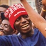 A young man in a red patterned cap smiling with his hand raised at a community event.