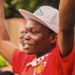 A young man in a white cap and red shirt smiling with his hand raised at a community event.